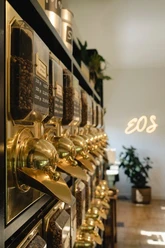 EOS1.jpg Reihe von goldenen Kaffeebehältern an einer Wand in einem eleganten Café mit Pflanzen im Hintergrund.