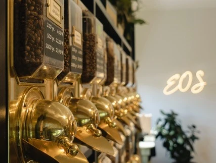EOS1.jpg Reihe von goldenen Kaffeebehältern an einer Wand in einem eleganten Café mit Pflanzen im Hintergrund.