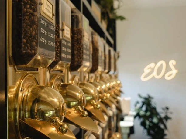 EOS1.jpg Reihe von goldenen Kaffeebehältern an einer Wand in einem eleganten Café mit Pflanzen im Hintergrund.