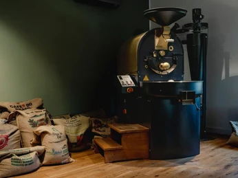 EOS2.jpg Industrieller Kaffeeröster in einem Raum mit Jutesäcken voll Kaffeebohnen auf Holzboden.