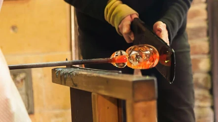Fertigungstechniken Eine Person formt mit Werkzeugen vorsichtig heißes Glas im Blasprozess auf einem Holztisch.