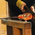 Probieren Sie es selbst.jpg Eine Person formt mit Werkzeugen vorsichtig heißes Glas im Blasprozess auf einem Holztisch.