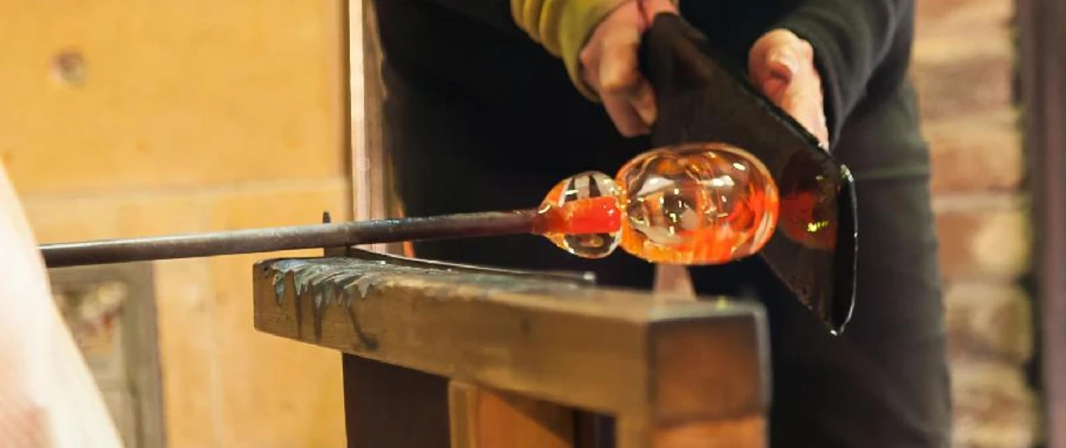Probieren Sie es selbst.jpg Eine Person formt mit Werkzeugen vorsichtig heißes Glas im Blasprozess auf einem Holztisch.