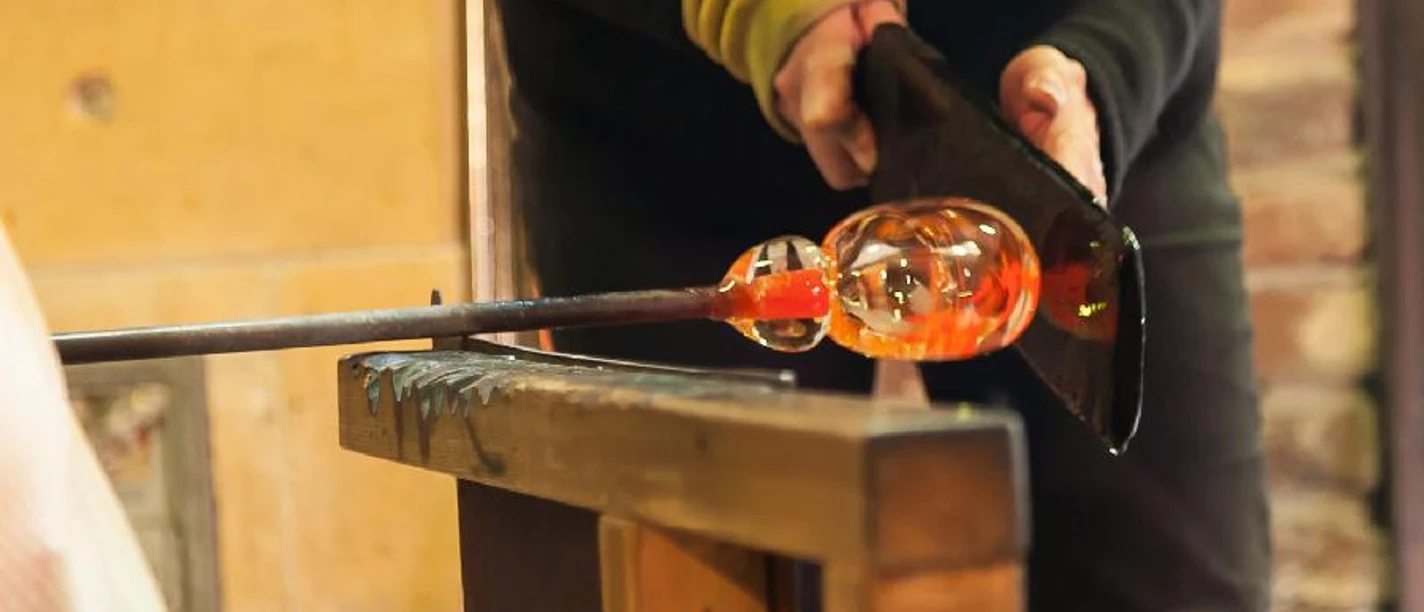 Probieren Sie es selbst.jpg Eine Person formt mit Werkzeugen vorsichtig heißes Glas im Blasprozess auf einem Holztisch.