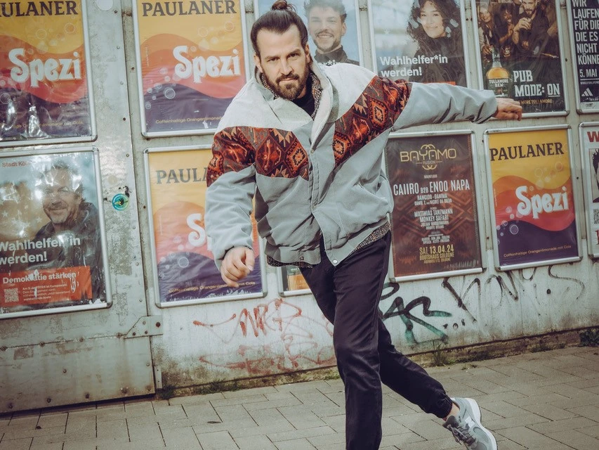 David Kebekus in lässiger Pose vor einer Plakatwand mit Zeitungstiteln und Getränkemarke.