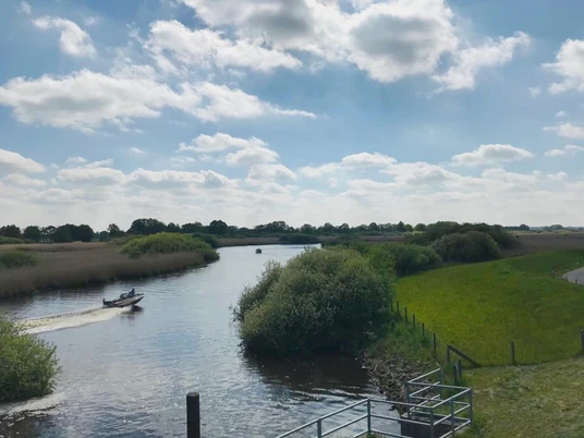 Blick auf die Soeste-Landschaftsfenster Tange Blick über die Soeste. Landschaftsfenster in Tange: Ein fließender Fluss mit saftig grünen Ufern, darauf ein Boot unter blauem Himmel mit lockeren Wolken.