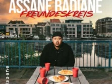 Assane Radiane sitzt an einem Tisch im Freien mit Essen und Getränken, im Hintergrund eine Wasserlandschaft.