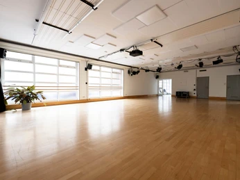 Un-Label Heller Tanzraum mit großen Fenstern, hellbraunem Holzboden, seitlichem Pflanzendekor und Deckenbeleuchtung.Bright dance room with large windows, light brown wooden floor, plant decor on the sides and ceiling lighting.
