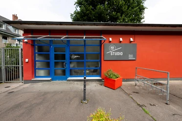 Un-label Das Bild zeigt die Vorderansicht des Un-label Studios mit einer markanten blauen Tür und großen Fenstern, flankiert von roten Wänden und einem Blumenkübel.