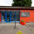 Un-label Das Bild zeigt die Vorderansicht des Un-label Studios mit einer markanten blauen Tür und großen Fenstern, flankiert von roten Wänden und einem Blumenkübel.The picture shows the front view of the Un-label studio with a striking blue door and large windows, flanked by red walls and a flower pot.