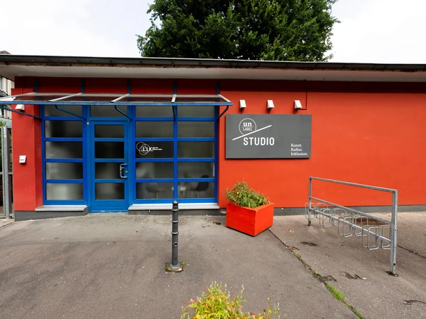 Un-label The picture shows the front view of the Un-label studio with a striking blue door and large windows, flanked by red walls and a flower pot.