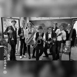 Eine Gruppe von acht Musiker*innen und einem Kind posieren lachend mit Instrumenten wie Saxophon und Gitarre.