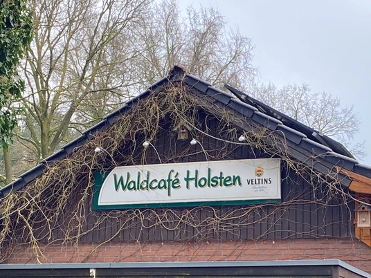 © Gemeinde Salzbergen Hausgiebel mit Schild „Waldcafé Holsten“ unter dunklem Dach, umrankt von Kletterpflanzen und Ästen