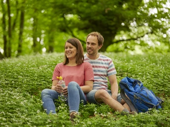 Zwei Personen sitzen lächelnd auf einer grünen Wiese im Wald, umgeben von blühendem Bärlauch.