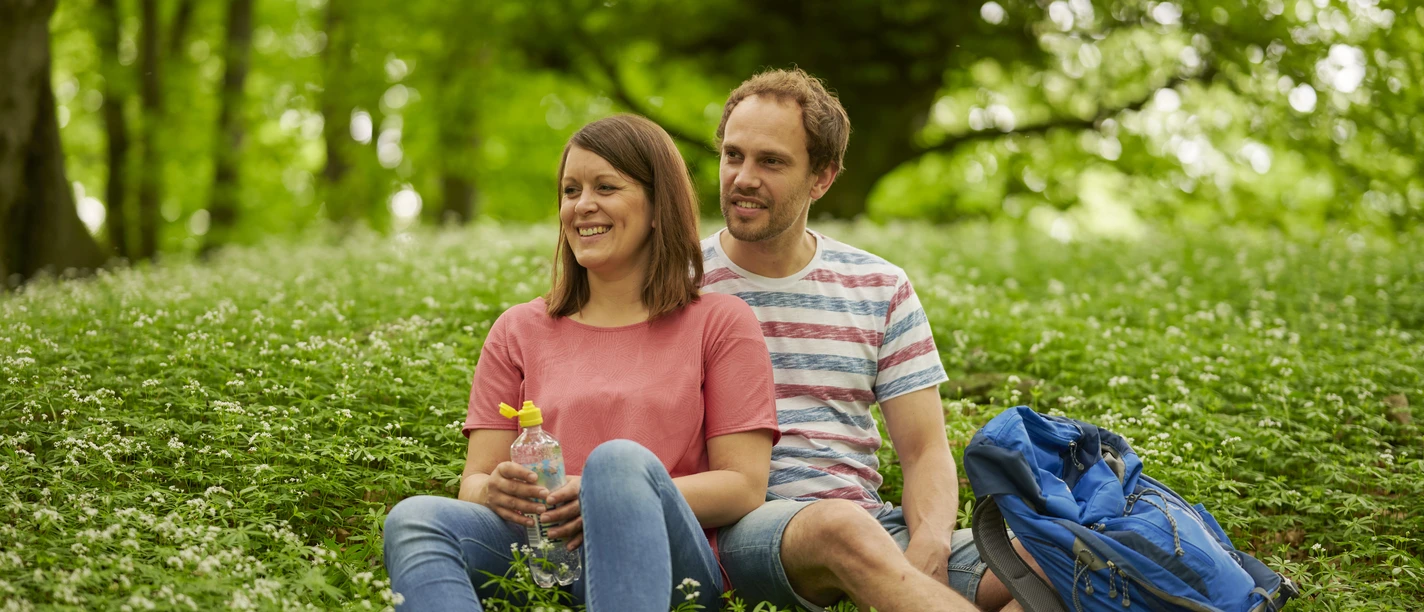 Wandern Rast Zwei Personen sitzen lächelnd auf einer grünen Wiese im Wald, umgeben von blühendem Bärlauch.