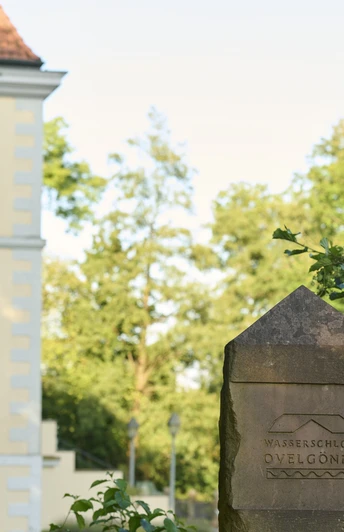 Das Foto zeigt einen Stein mit der Inschrift "Wasserschloss Ovelgönne" vor einem historischen Gebäude.