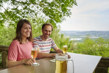 Wilder Schmied Paar genießt Bier auf Terrasse mit Blick über grüne Landschaft bis zum weitläufigen Tal.