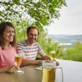 Wilder Schmied Paar genießt Bier auf Terrasse mit Blick über grüne Landschaft bis zum weitläufigen Tal.