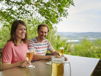 Wilder Schmied Paar genießt Bier auf Terrasse mit Blick über grüne Landschaft bis zum weitläufigen Tal.