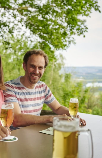 Wilder Schmied Paar genießt Bier auf Terrasse mit Blick über grüne Landschaft bis zum weitläufigen Tal.