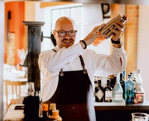 Mann mit Brille und Schürze mixt lächelnd einen Cocktail vor einer Bar mit diversen Getränke.