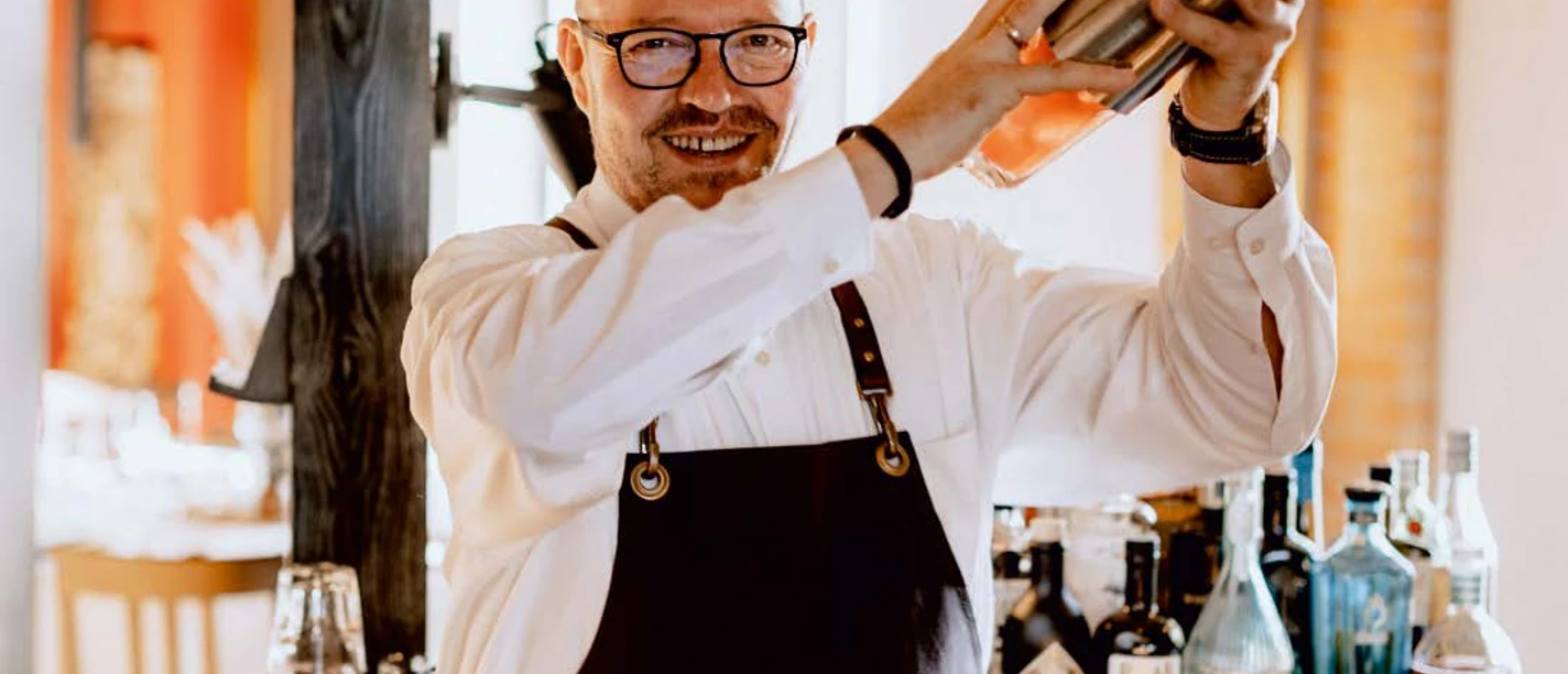 Mann mit Brille und Schürze mixt lächelnd einen Cocktail vor einer Bar mit diversen Getränke.