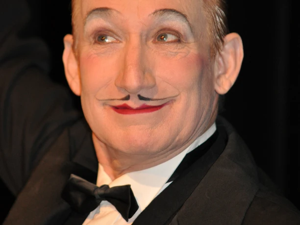 DSC_0133.jpg Ein elegant gekleideter älterer Mann im Smoking mit kunstvoll gemaltem Gesichtsausdruck und einem humorvollen, schelmischen Lächeln. Ein weißes Blüten-Accessoire ziert sein Revers.