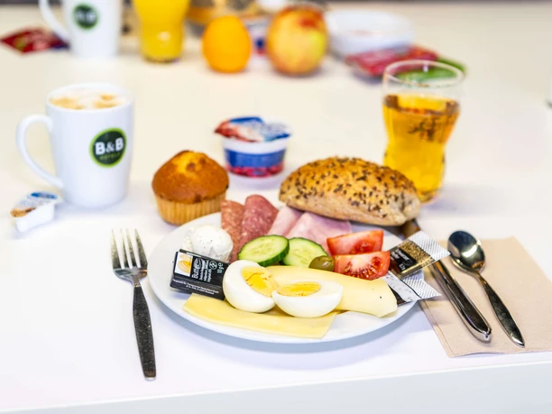 Frühstück im B&B HOTEL Düsseldorf-Hilden Ein Teller mit frischem Frühstück: Aufschnitt, belegtes Brötchen, Heißgetränk und Saft im Hintergrund.