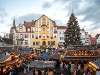 Weihnachtsmarkt mit festlich beleuchteten Buden, bunten Lichtern und einem großen geschmückten Tannenbaum.