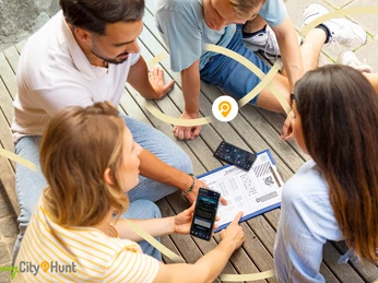 Herford-CityHunt-CityHunters GmbH & Co.KG_3.jpg Vier Personen sitzen auf Holzboden und betrachten ein Rätsel auf Papier; Handys in der Hand.