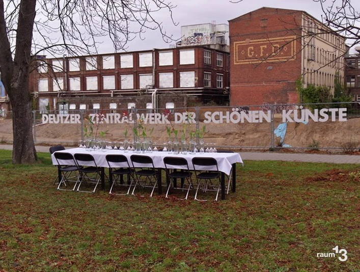 raum13 Eine lange, festlich gedeckte Tafel steht unter freiem Himmel vor einer alten Backsteinfabrik.A long, festively laid table stands in the open air in front of an old brick factory.
