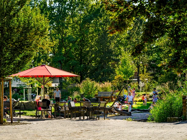 Flora-Westfalica-Park_Spielerei4_Foto M Wallenfang.jpg Menschen entspannen an einem sonnigen Tag in einem grünen Park mit roten Sonnenschirmen und Sitzbereichen.