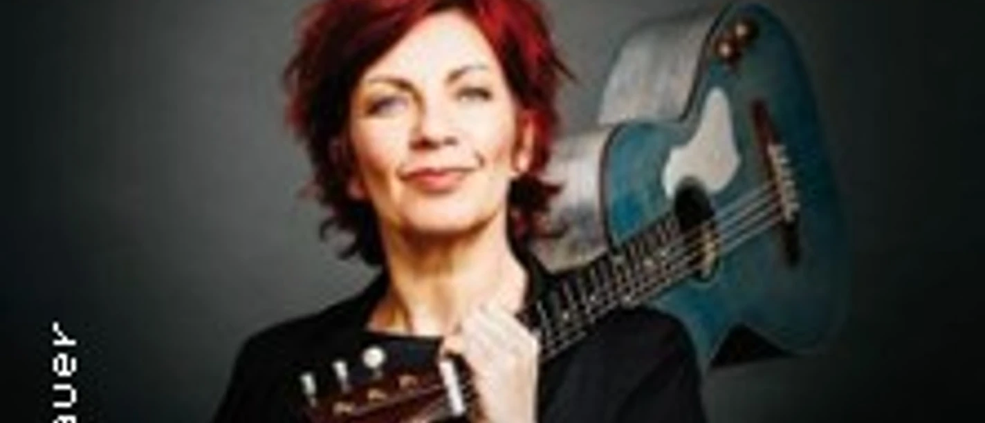 Eine Frau mit roten Haaren posiert lächelnd vor einer neutralen Wand und hält eine Gitarre im Arm.