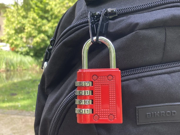 IMG_1081.jpg Ein schwarzer Rucksack auf einer Bank, gesichert mit einem roten Vorhängeschloss, im Freien.