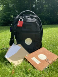 IMG_1083.jpg Ein schwarzer Rucksack mit Notizbuch und Papier liegt auf einer grünen Wiese unter sonnigem Himmel.