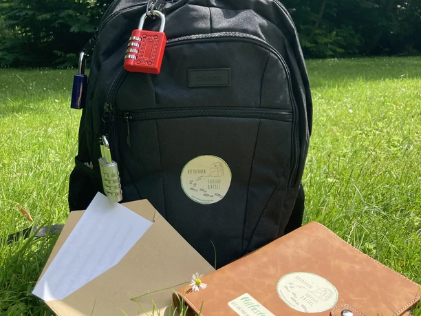 IMG_1083.jpg Ein schwarzer Rucksack mit Notizbuch und Papier liegt auf einer grünen Wiese unter sonnigem Himmel.
