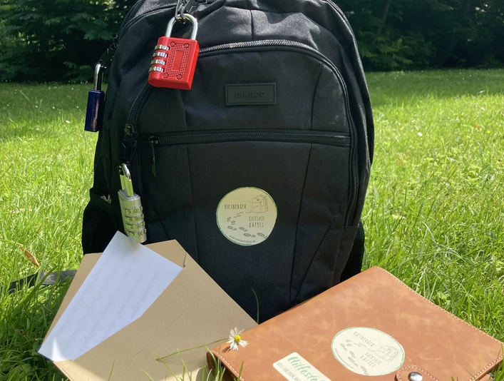 IMG_1083.jpg Ein schwarzer Rucksack mit Notizbuch und Papier liegt auf einer grünen Wiese unter sonnigem Himmel.