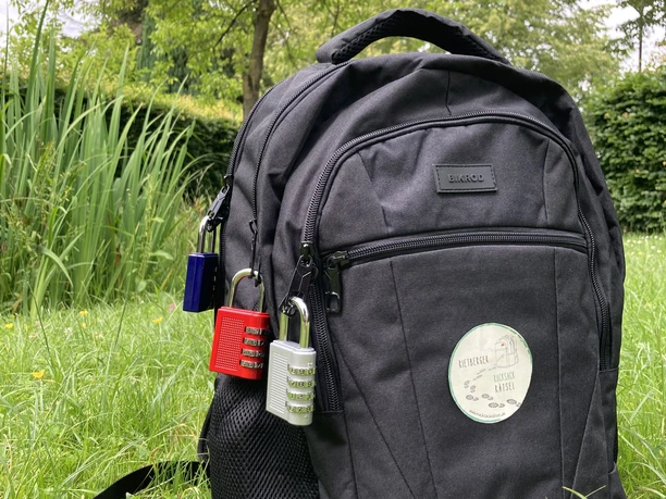 IMG_1098.jpg Ein schwarzer Rucksack steht auf einer grünen Wiese, dekoriert mit bunten Vorhängeschlössern.