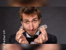 Ein Mann im Anzug mit verschmitztem Lächeln präsentiert Münzen in beiden Händen vor einem dunklen Hintergrund.