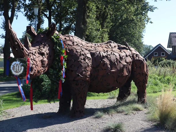 Nashorn-Spohle-DerWiefelsteder.JPG Das Bild zeigt eine lebensgroße Statue eines Nashorns in einer ländlichen Umgebung. Die Statue besteht aus rostigem Metall und trägt einen Kranz um den Hals, was auf eine Feier oder ein lokales Event hindeutet. Im Hintergrund sind Bäume und ein Wohnhaus zu sehen.