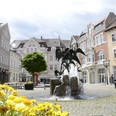 Brunnen mit Skulpturen in belebtem Platz; historische Gebäude umgeben die Szene, Blumen im Vordergrund.