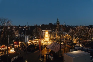 Ein festlicher Weihnachtsmarkt bei Nacht, beleuchtet von zahlreichen Lichtern. Menschen schlendern zwischen den Ständen und genießen die weihnachtliche Atmosphäre.