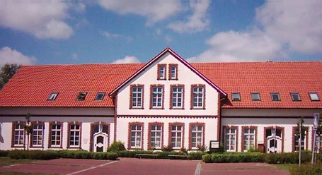 RathausKirchstraße10.jpg Das Bild zeigt ein zweistöckiges Gebäude mit leuchtend roten Dachziegeln und weißen Wänden. Die Fassade hat mehrere symmetrisch angeordnete Fenster mit roten Backsteinrahmen.