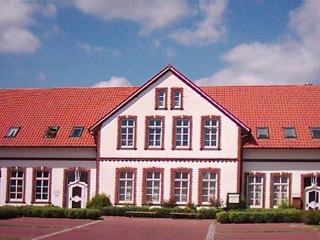 RathausKirchstraße10.jpg Das Bild zeigt ein zweistöckiges Gebäude mit leuchtend roten Dachziegeln und weißen Wänden. Die Fassade hat mehrere symmetrisch angeordnete Fenster mit roten Backsteinrahmen.