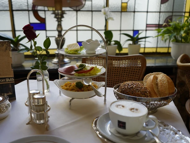 Cafe Wahlen Schön hergerichteter Frühstückstisch mit Brötchen, Cappuccino, Etagere und Vase mit Rose.