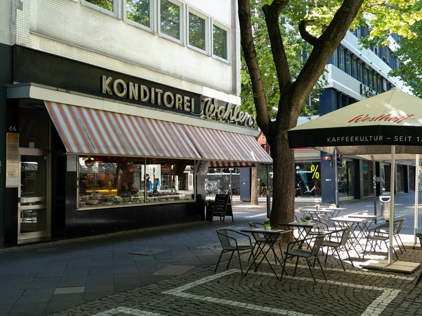 Cafe Wahlen Außenansicht vom Cafe mit orange-weiß gestreifter Markise. Unter einem Sonnenschirm laden Tische und Stühle zum verweilen ein.