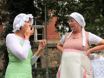 Die Waschweiber der Äbtissin Elisabeth 03.jpg Zwei Frauen in traditioneller Kleidung mit Hauben unterhalten sich lebhaft auf einer historischen Straße.