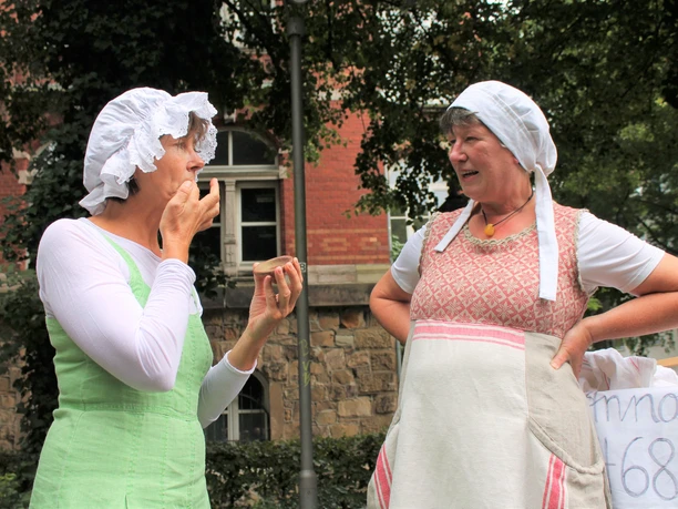 Die Waschweiber der Äbtissin Elisabeth 03.jpg Zwei Frauen in traditioneller Kleidung mit Hauben unterhalten sich lebhaft auf einer historischen Straße.