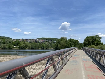 Fuß- und Radbrücke.jpg Eine moderne Fuß- und Radbrücke überspannt einen Fluss, umgeben von grüner Natur und hügeliger Landschaft.