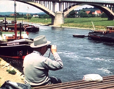 Ein Mann mit Fernglas beobachtet Schiffe auf der Weser, während eine beeindruckende Brücke im Hintergrund das Flussufer überquert.